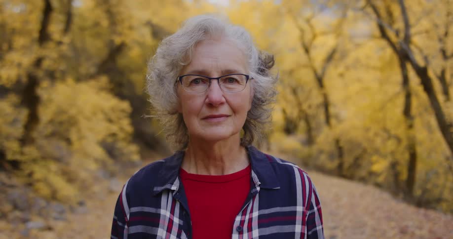 An elderly woman is standing on a dirt road and there are trees with fall colored leaves behind her