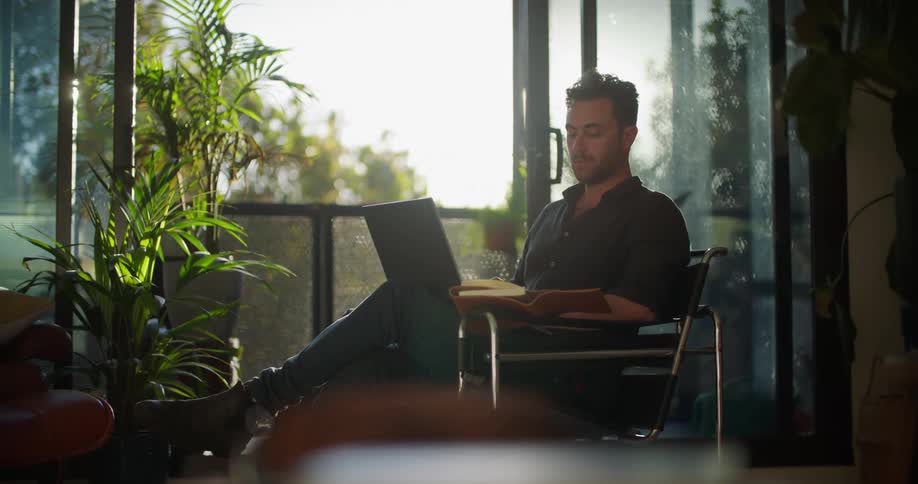 Man sitting in chair looks at book and types on laptop in living room