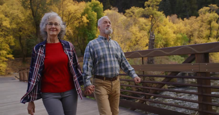 An elderly couple are outside holding hands as they walk across a footbridge and there is a forest of trees in bright fall colors behind them
