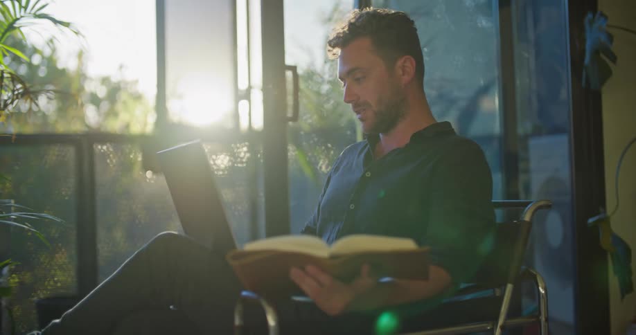 Man with blue shirt uses laptop making a video call and book in hand