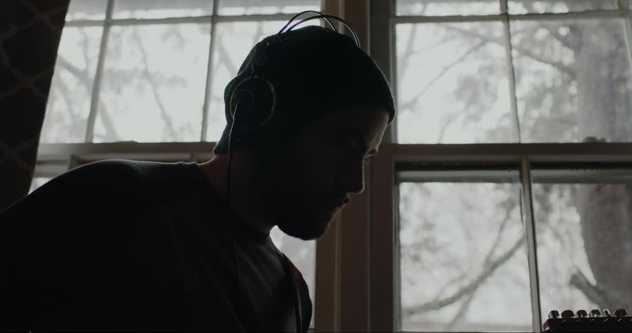 A man wearing headphones plays his guitar near a large window