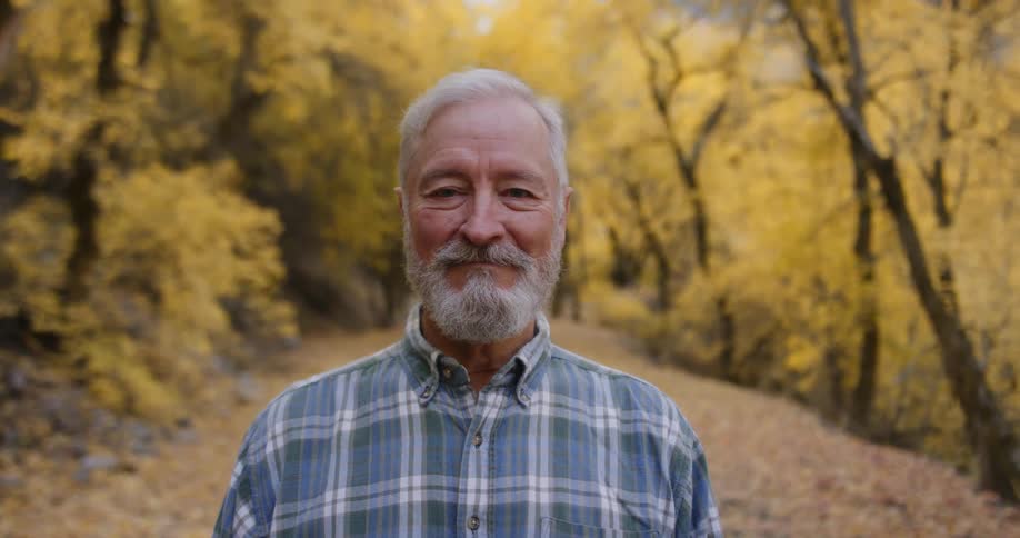 An elderly man smiles into the camera while standing on a path through a woodland during the day