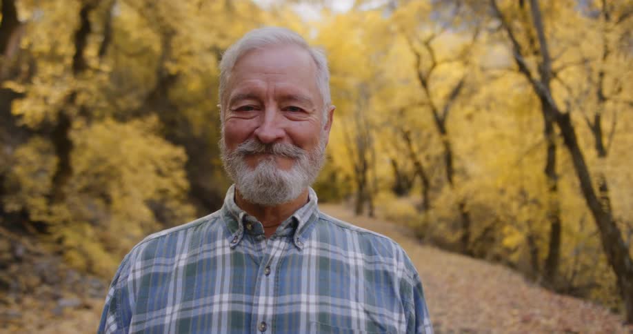 An elderly man stands on a path in a forest filled with fall tree and smiles for a photo