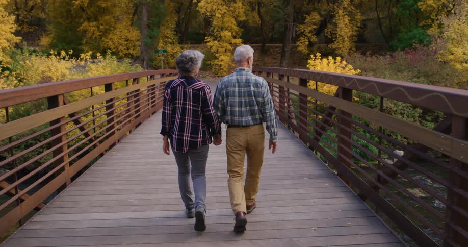 Elderly couple hold hands while walking on a bridge over a river near the mountains