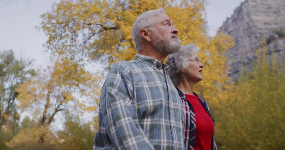 An elderly couple are walking by trees in fall colors and mountains are in the distance