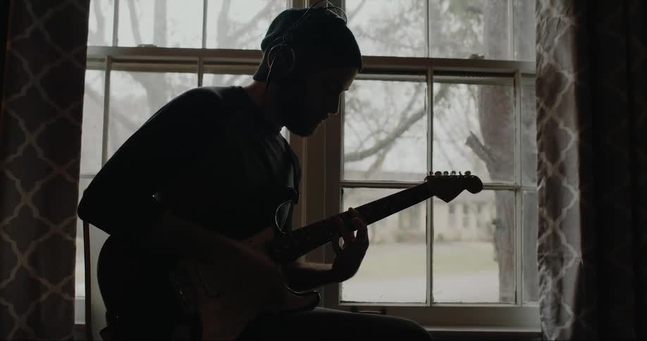 Silhouette of a young man playing guitar near a window at home