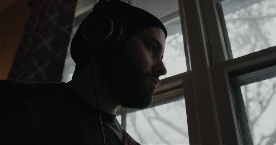 A man is near a window wearing headphones and moving his head to the music