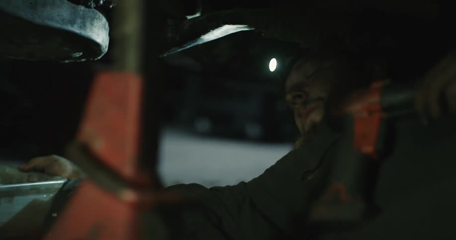 A mechanic lies under a car and pulls a container towards him and then loosens bolts before doing an oil change