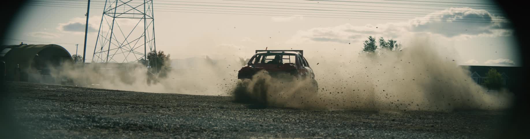 Driver maneuvers crinkled demo derby car in circle spewing dirt with building and other vehicles in the background