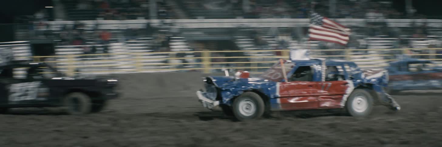 Audience watches demo cars drive along dirt arena and crash into each other at night