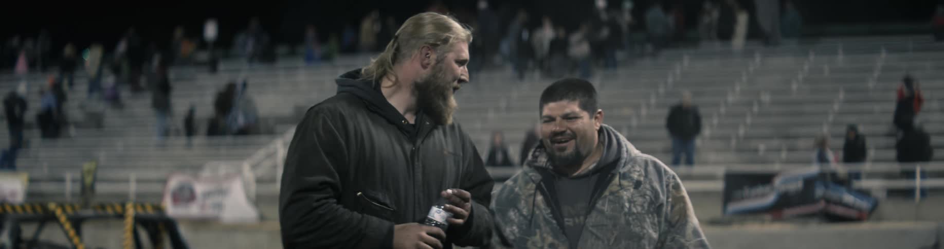 A man takes a drink of water and talk to another man in a stadium after a car race and then they bumps fists