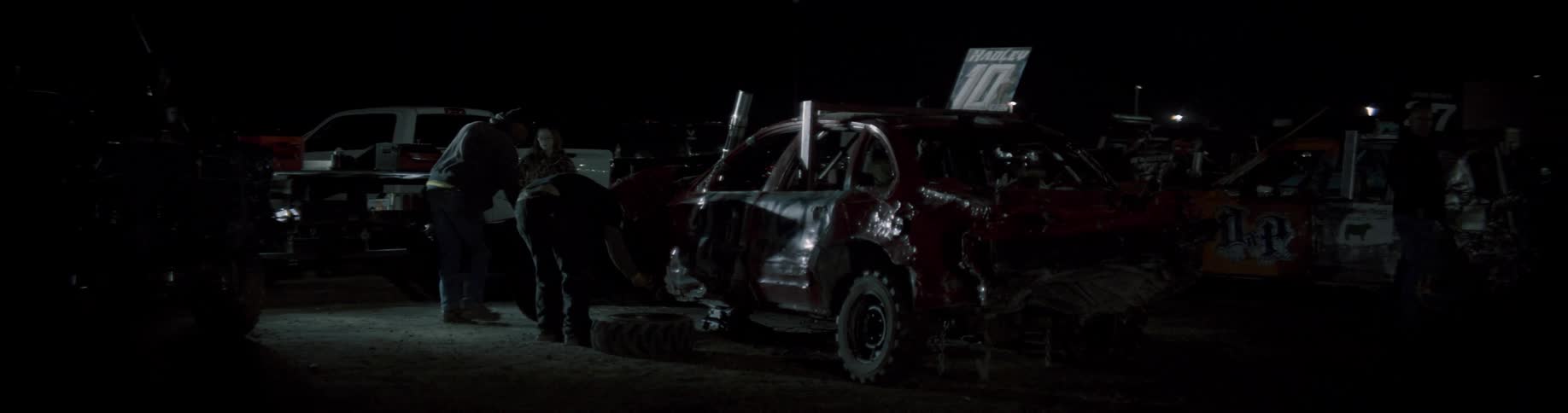 Two people are standing next to a beat up race car with a tire off