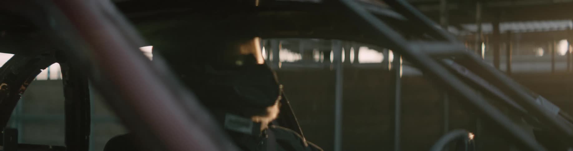 The driver in a demolition derby car is wearing a helmet and goggles and the sun shines brightly in the distance