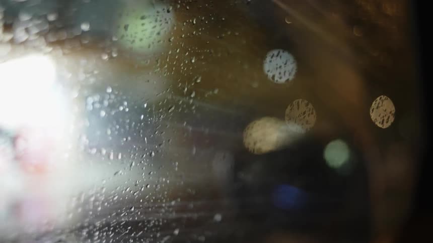 Vehicle drives past businesses with lights that are turned into glare by water on side window