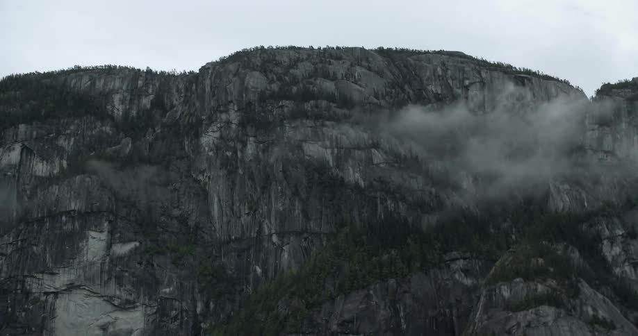 Clouds move past a sheer mountain cliff with trees along the ridge on a gray day