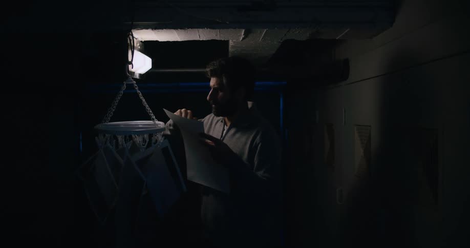 Male photographer stands in darkroom wiping photograph near other hanging photographs