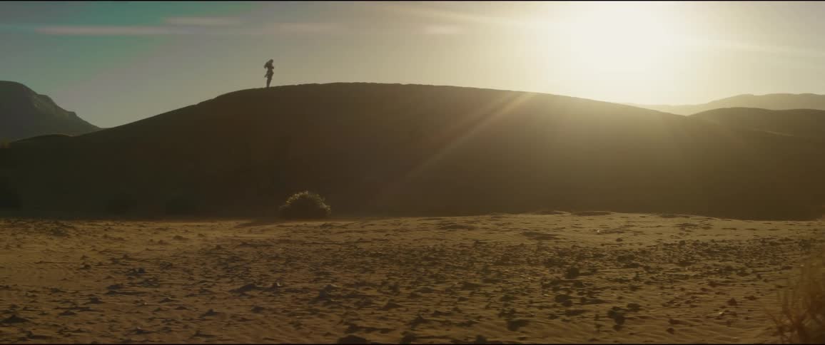 Silhouette of a person standing on top of a hill in the desert as the sun shines brightly in the sky