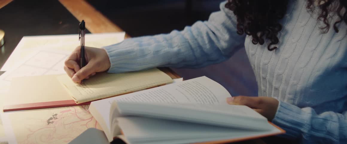 Girl with curly hair and turquoise sweater sits at desk writing down in notebook in the library