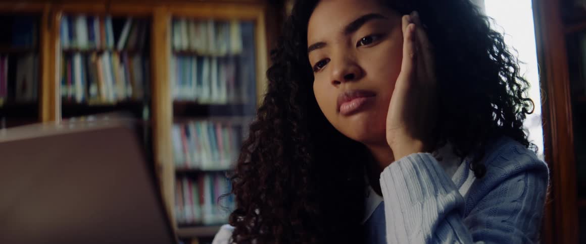 A teenage girl sits in a library and rubs her head as she works on her laptop