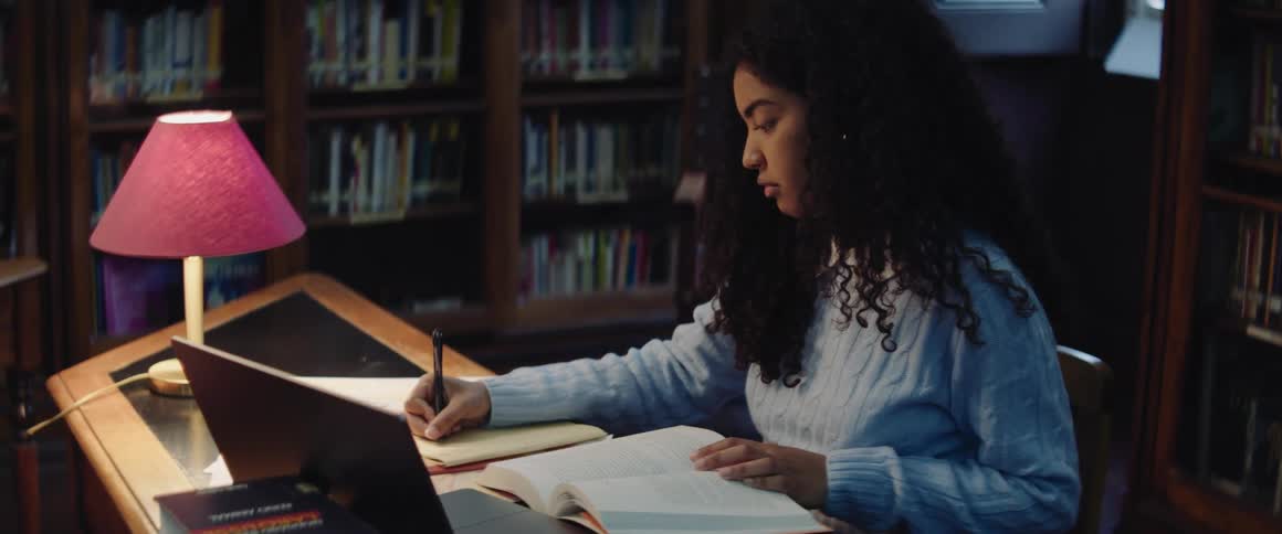 A teenage girl is sitting at a table in a library taking notes from an open book in front of her