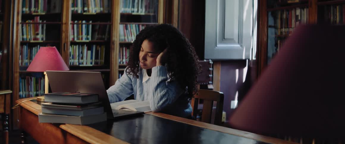 A teenage girl is sitting at a table in a library with a book in front of her and she is using a laptop computer