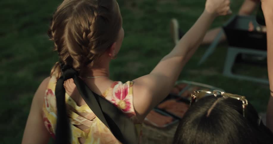A woman clinks beer bottles with others as they sit outside grilling and drinking