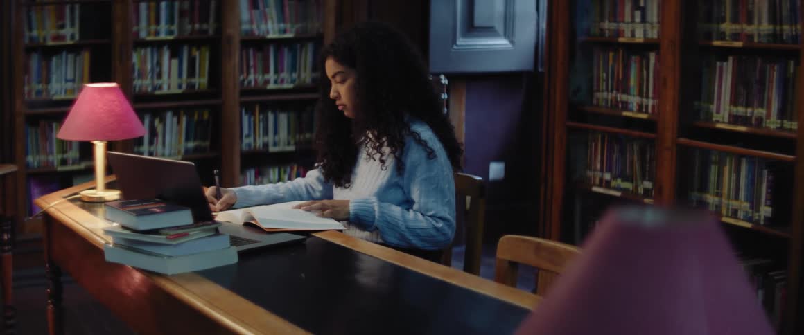 A teenage girl is writing at a library table with a stack of books and a laptop computer