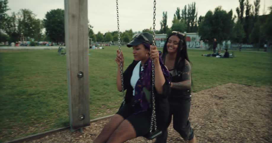 Friends run to swingset and one swings while the other pushes as other park visitors have picnics in the background