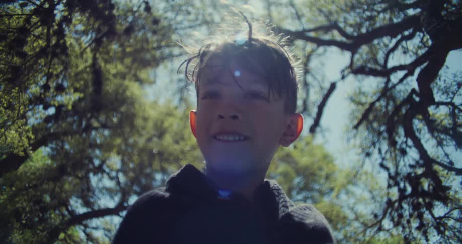 A young boy smiles as he stands outside near trees and the sun is shining brightly in the sky