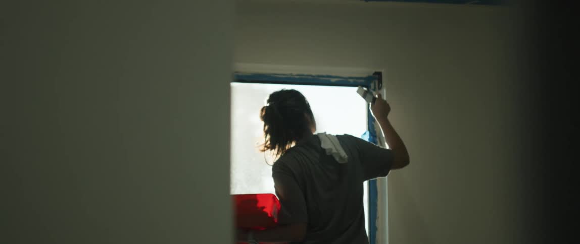 A woman with a pony tail stands in front an open window holding an orange paint tray on her left arm dabbing white paint on the top right hand corner of the window frame