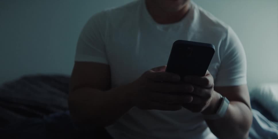 A man wearing a white tshirt is holding a cell phone and using both hands