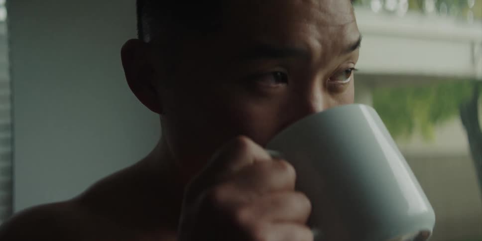 A shirtless man drinks from a coffee mug while gazing out of a window during the day
