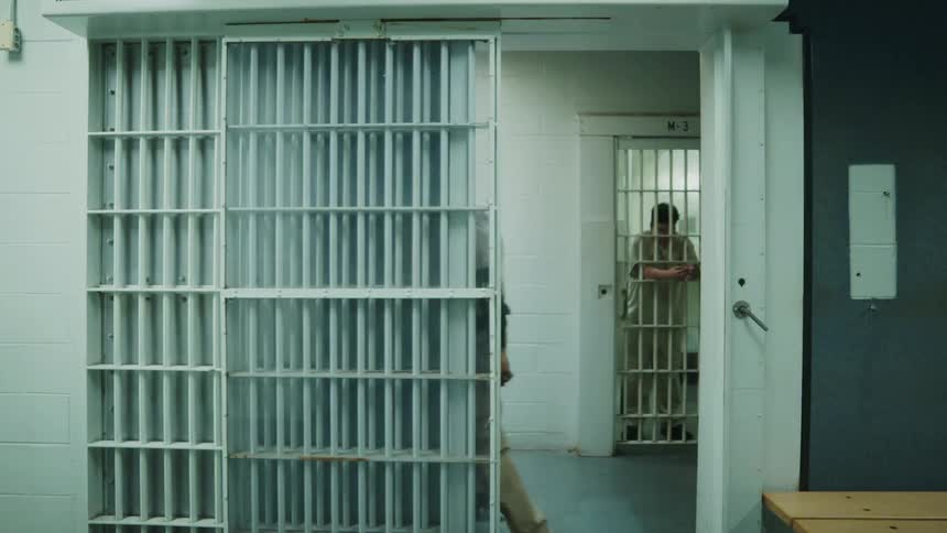 A man is leaning on the bars of a jail cell and an officer brings in another inmate