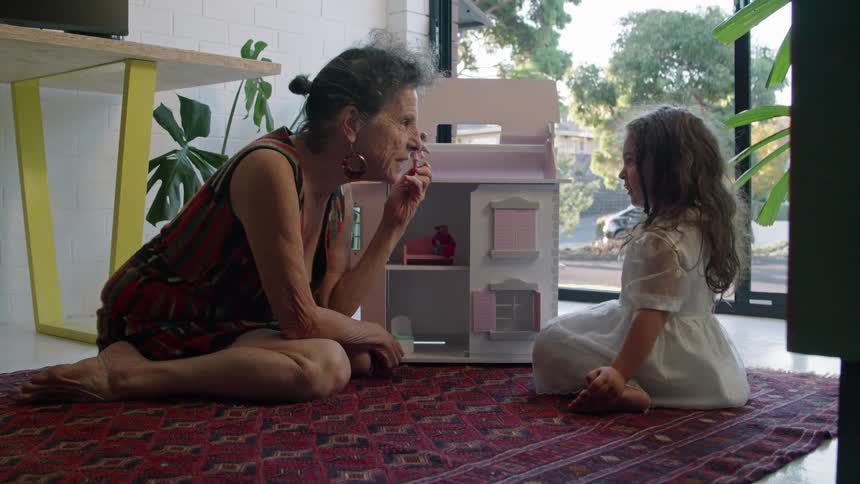 A woman is sitting on the floor playing with dolls and a doll house with her granddaughter