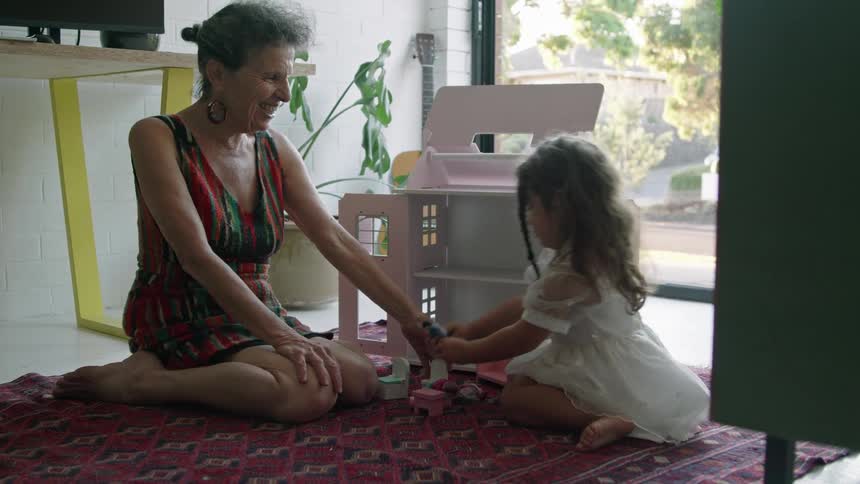 A woman sits on the floor and plays with her granddaughter in front of a doll house