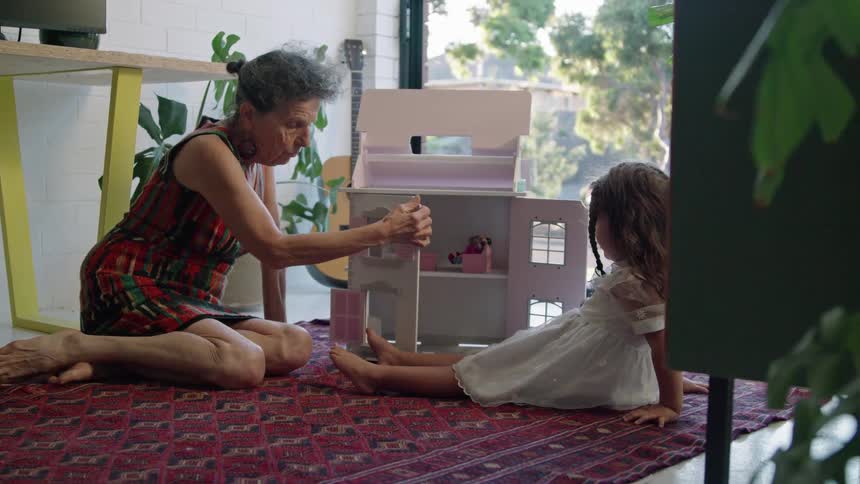 A woman is sitting on the floor playing with a doll house with her granddaughter