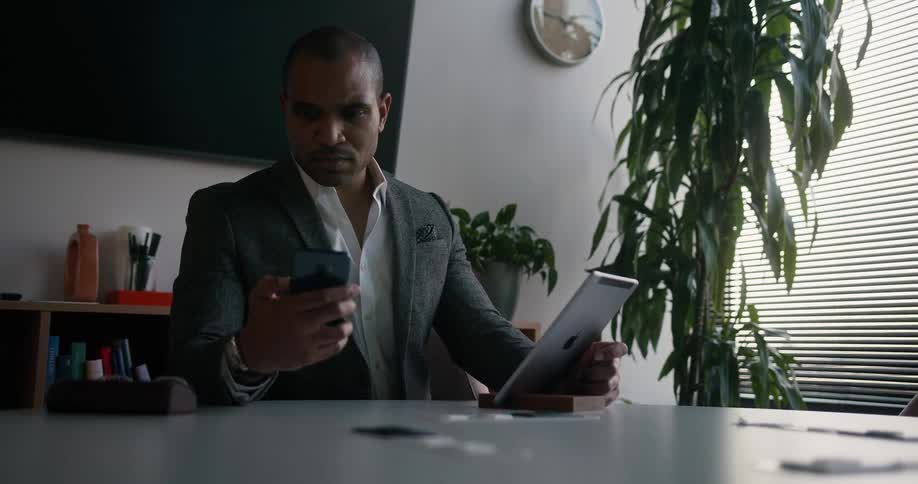 A man is sitting at a table holding a tablet computer and he answers his phone