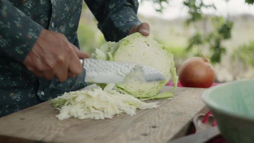 A man cuts strips of cabbage on a cutting board outdoors