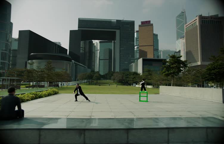 A man and woman walk and sit down to watch a man and child perform in a square in front of office buildings