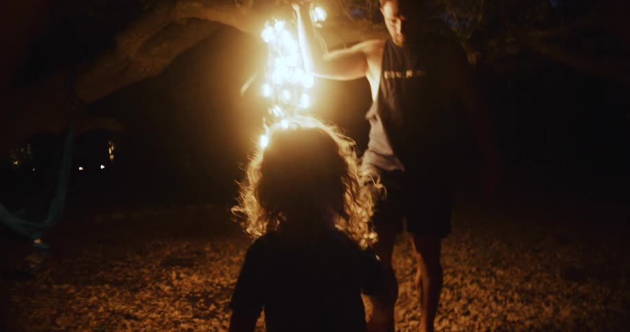 A child walks toward dad who is carrying a glowing string of lights in the backyard