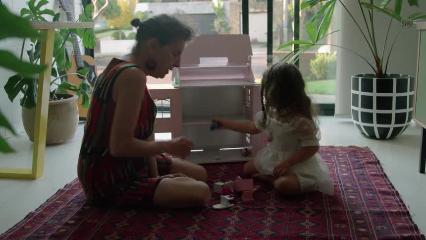 Old woman with striped dress plays with toddler girl with wavy hair and white dress next to pink model house sitting on red carpet in living room at home