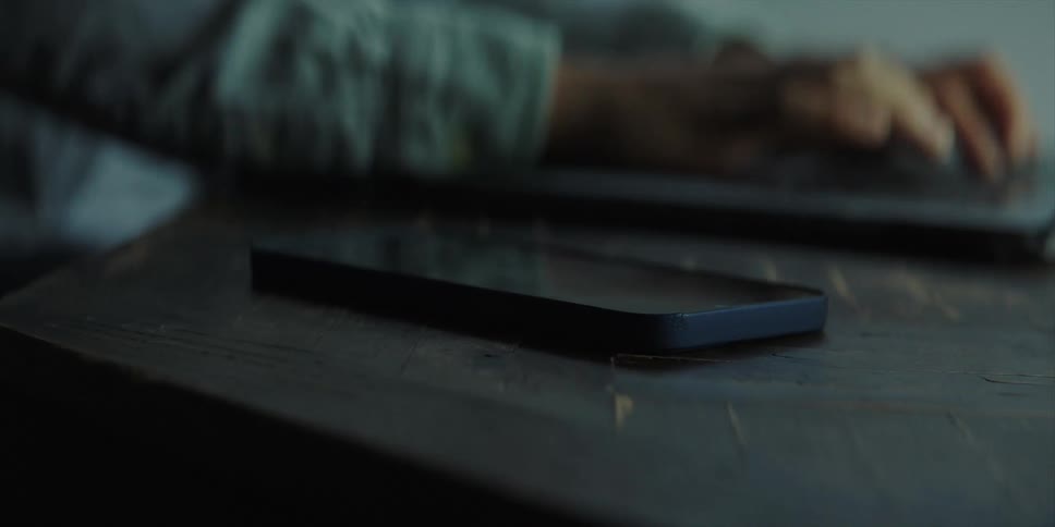 Man with blue shirt types on laptop and picks up smartphone sitting at table