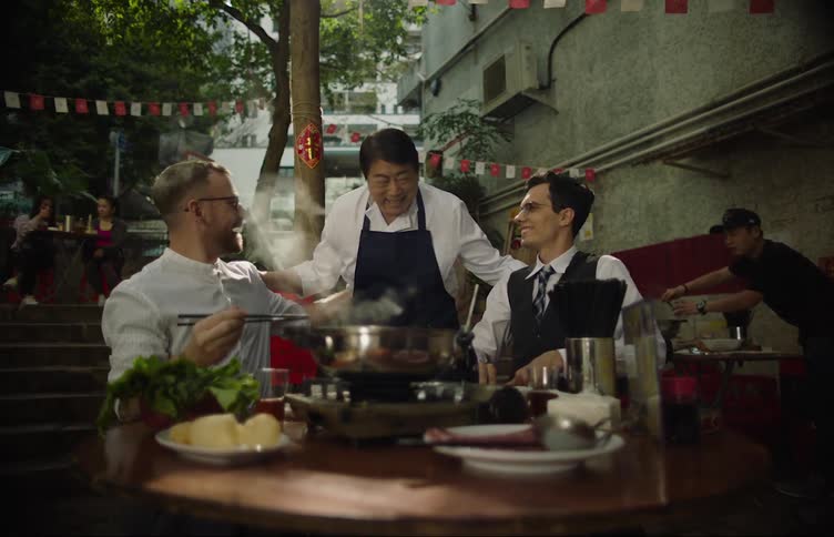 A waiter drops off hot pot vegetables and greets the two men sitting at the outdoor dining table