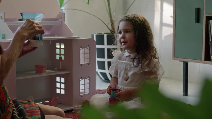 Old woman with striped dress plays with toddler girl with wavy hair and white dress next to pink model house sitting on red carpet in living room at home