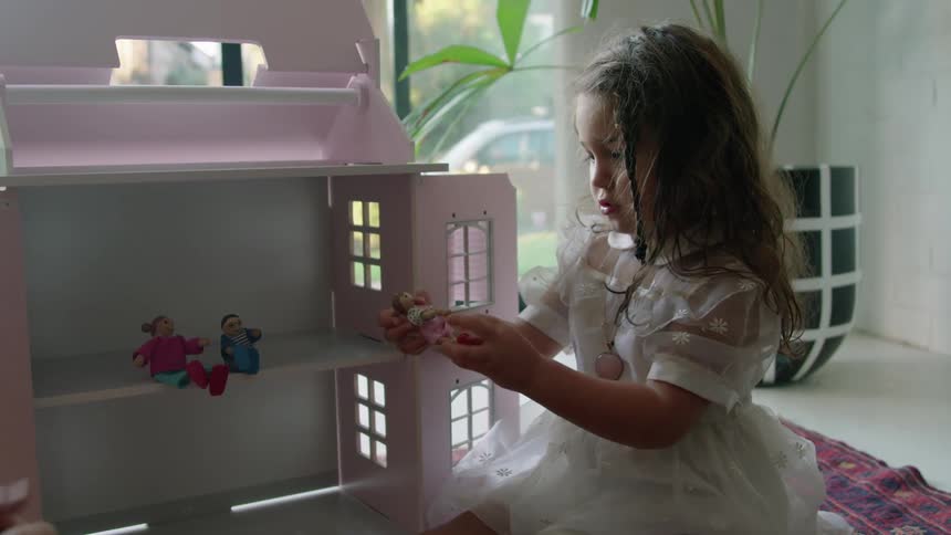 Old woman with striped dress plays with toddler girl with wavy hair and white dress next to pink model house sitting on red carpet in living room at home