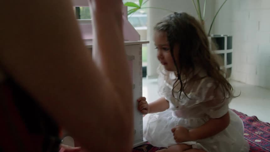 Old woman with striped dress plays with toddler girl with wavy hair and white dress next to pink model house sitting on red carpet in living room at home
