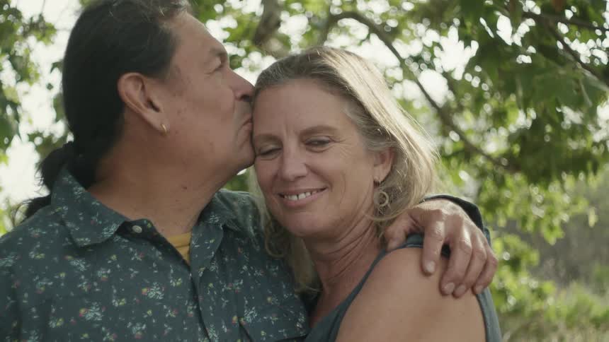 A man and woman are hugging and he kisses her forehead as they stand under a tree