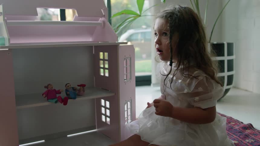 A little girl wearing a white dress is putting dolls in her doll house