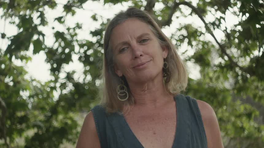 Happy blonde middle aged woman standing next to a tree outdoors on a cloudy day