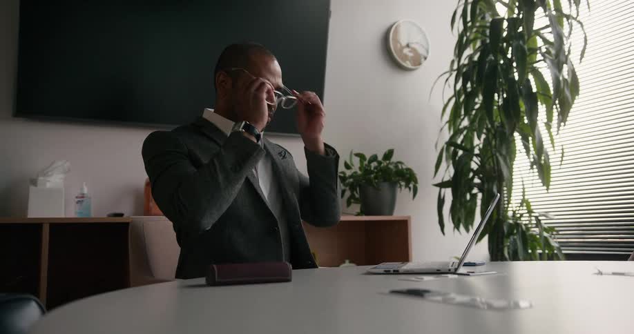 A man is sitting at a table and he puts on his glasses and types on a laptop computer